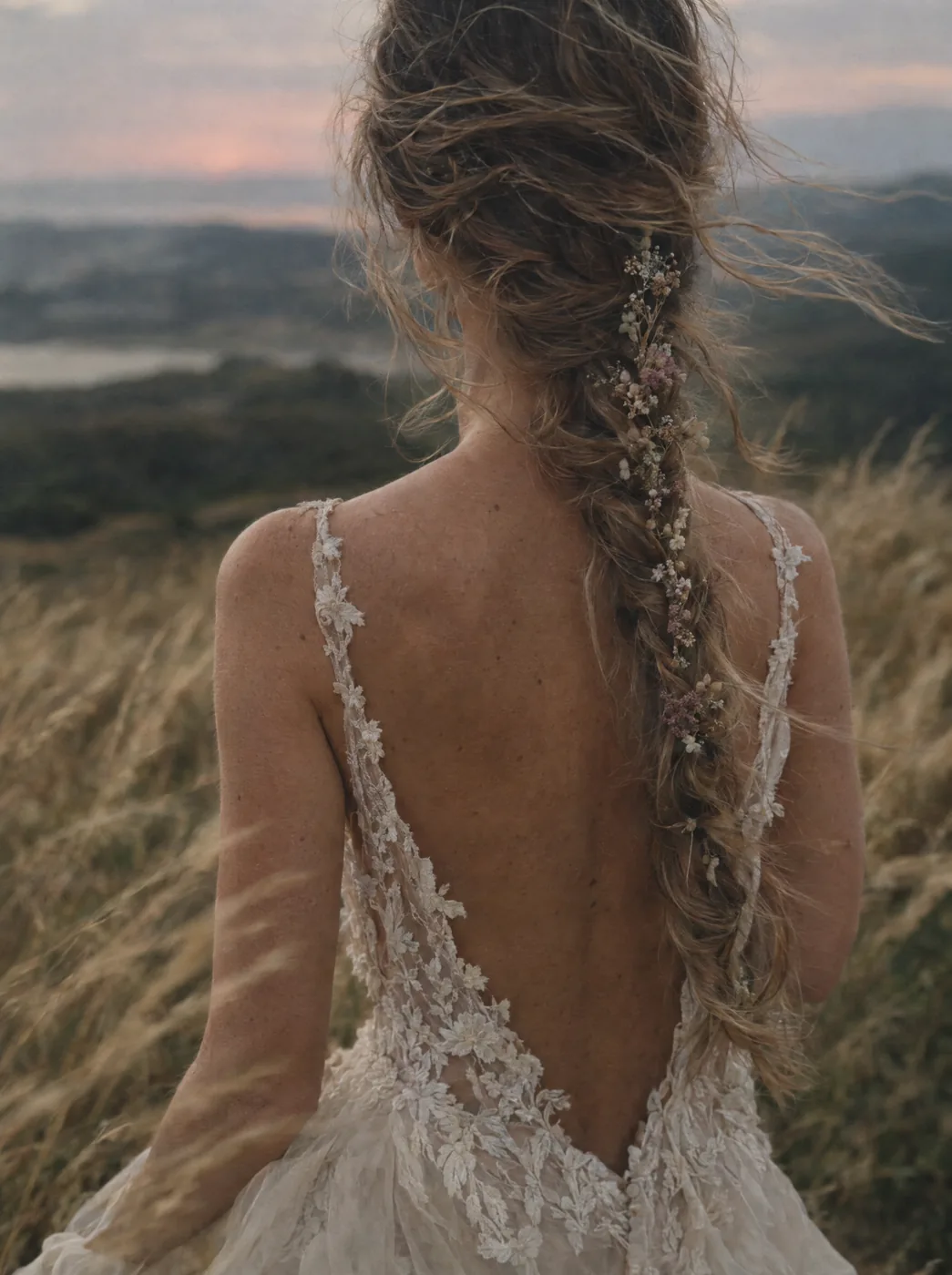 Una sposa di spalle nell'erba alta al tramonto, la treccia intrecciata di fiori secchi e la schiena in pizzo